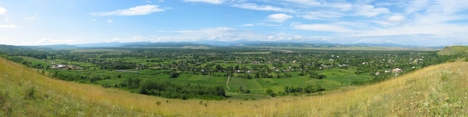 A view from a mountainside onto a village located in a mountain valley. Summer panoramic landscape. Village houses in the distance. Kaladzhinskaya village, Krasnodar Krai, Russia. Wide panorama.