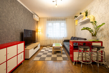 Apartment interior. Living room with furniture: dark gray sofa, red chest of drawers, metal chairs, wooden coffee table, shelves with indoor plants, TV. The window is covered with a tulle curtain.