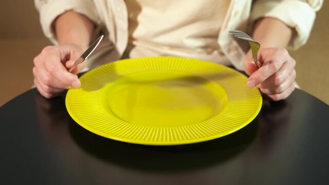 Person waiting for food at a table with an empty plate. Anonymous person sitting at a restaurant table, holding a fork and knife over an empty yellow plate, nervously waiting for their meal