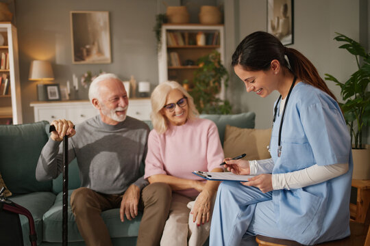 Home health nurse sits with a senior couple in their living room, discussing care plans while taking notes on a clipboard during a home visit.