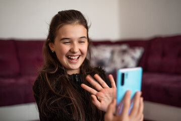 A happy teenage girl waving hand greeting gesture hello on video call while using a smartphone at home.
