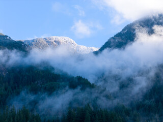 Fototapeta premium Snow-Capped Mountain Ridge Over Misty Forest Valley Under Clear Blue Sky in Howe Sound