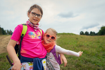 Older Sister in Bright Pink Hiking Shirt Hugging Younger Sister with Orange Scarf and Backpack on Grassy Countryside Trail