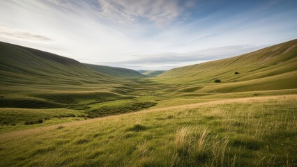 Fototapeta premium Wide landscape shot of a peaceful green valley showing rolling hills, meadows, and a bright partly cloudy sky.