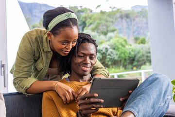 Fototapeta premium African American couple sitting on dark upholstered sofa near glass door, holding tablet together