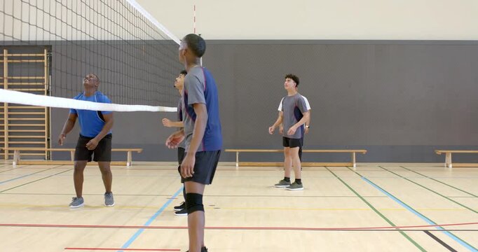 Diverse male teammates spiking volleyball over net after opponent serve to score point at gym