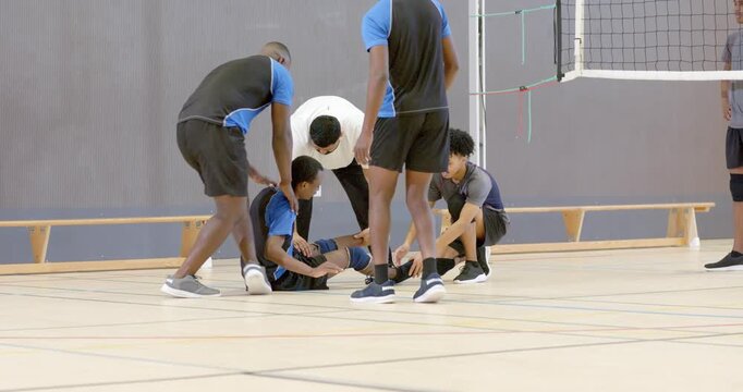 Asian man collapsing onto gym floor near net, teammates and coach rushing in, examining injured leg
