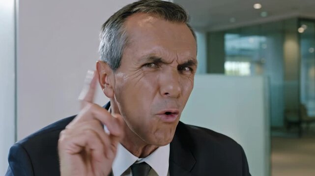 A mature businessman in a modern office lobby is shaking his finger and head, saying "No" with a stern gesture to express disapproval