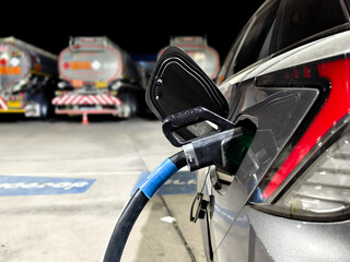 Naklejka premium Electric car charging with fuel tanker trucks in background