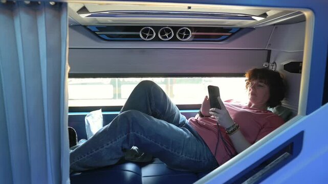 Sleeper Bus Teen Reclining In Car Seat Watching Digital Content On Mobile Device. Young Passenger Lounging Comfortably In Sleeper Seat Viewing Videos On Smartphone During Scenic Road Trip
