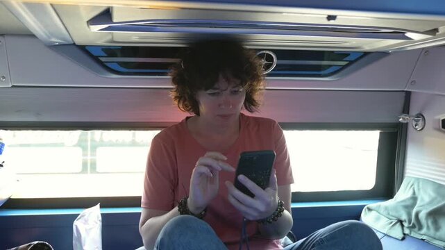 Sleeper Bus Backpacker In Sleeper Bus Bunk Scrolling Phone, Quiet Daylight Scene Showing Overhead Vent, Window Light, Loose Jacket, Earbuds And Backpack, Reflective Mood Of LongDistance