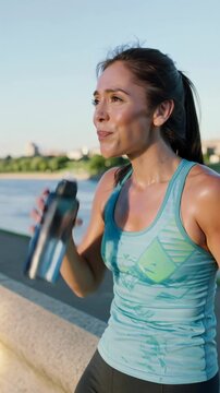 Young woman drinking water from a bottle after outdoor exercise by the riverside, vertical shot