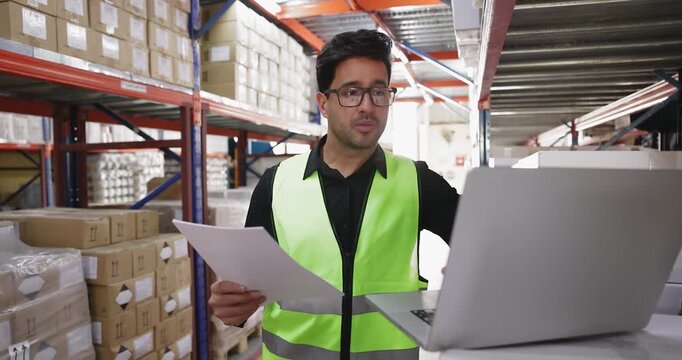 Serious Latin male storage supervisor counting goods after delivery, making inventory, using laptop at metal shelves, looking at boxes, consulting paper waybill and computer