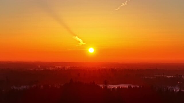 Drone view of the sun still above the horizon during sunset over a snowy winter landscape in Amatciems, Latvia. Warm orange light fills the sky, creating a calm and uplifting winter scene.