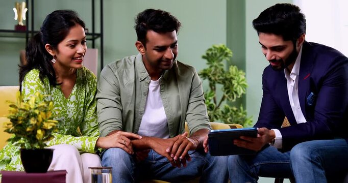 Indian couple discussing with agent at home with tablet computer during financial investment meeting with bank representative reviewing documents while sitting on sofa in cozy modern living room inter