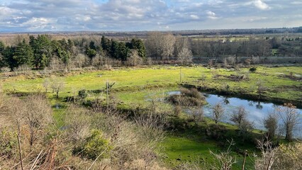 Wide landscape with green fields, a pond, and a forest on a cloudy day.