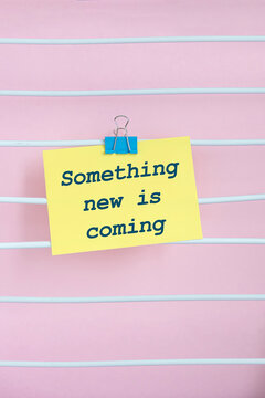 A yellow sticky note with the text 'Something new is coming' attached to a pink striped background. The note is clipped with a blue paperclip.
