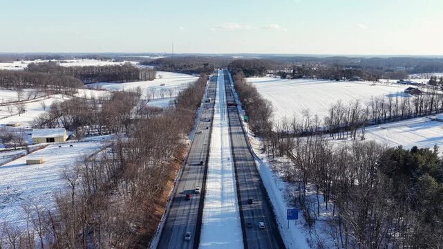 Aerial view of long rural highway cutting through snow-covered farmland in America, with light traffic driving between frozen fields and bare winter trees under clear daylight. Wide shot. PA MD border