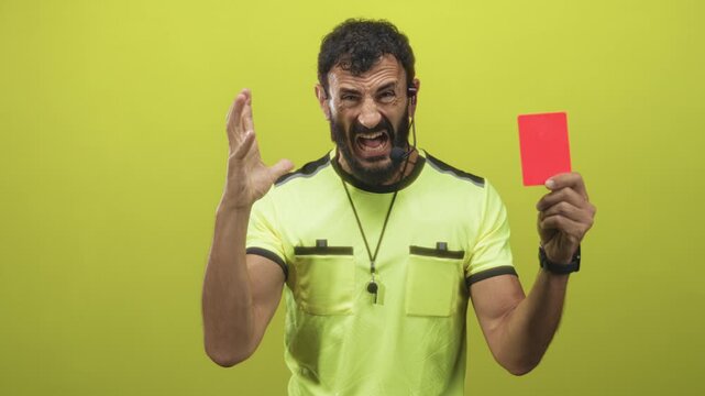 Referee man with beard holds red card in hand, grimaces and raises other hand while wearing whistle and headset in studio; discipline frustration.