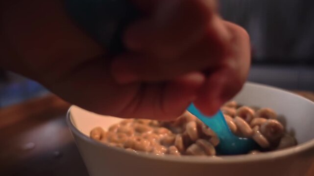 Medium close-up slow motion shot of a blue spoon lifting cereal and milk above a bowl, with milk dripping back into the bowl, highlighting texture, gravity, and motion during breakfast preparation.