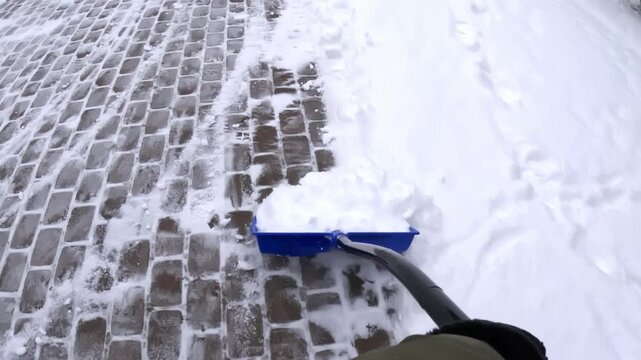POV pushing a blue snow shovel diagonally across cobblestone pavers, sweeping leftover powder into a rounded pile and finishing a driveway cleanup after snowfall.