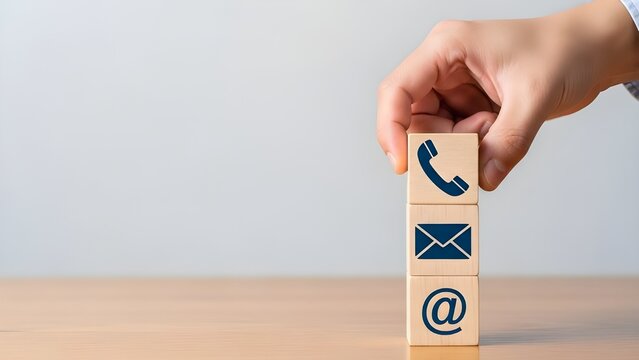 A hand stacking wooden blocks with contact icons like phone, email, and at symbol on a wooden table.