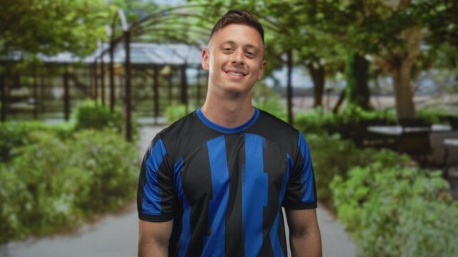Man wearing blue and black soccer jersey smiles and tilts head on a tree lined street; confidence team spirit.