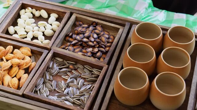 Wooden tray neatly divided into compartments: pistachios, watermelon seeds, apricot kernels, and sunflower seeds for snacking. Six matching terracotta cups waiting to be filled. 