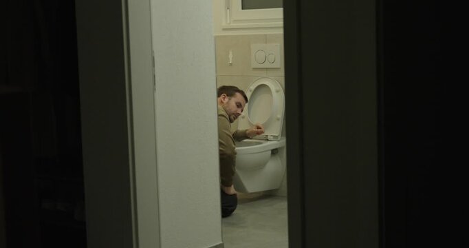 Nighttime scene of a man feeling nauseous while sitting on the toilet, viewed through a doorway. Concept of sickness, digestive issues, nighttime illness, and physical discomfort.