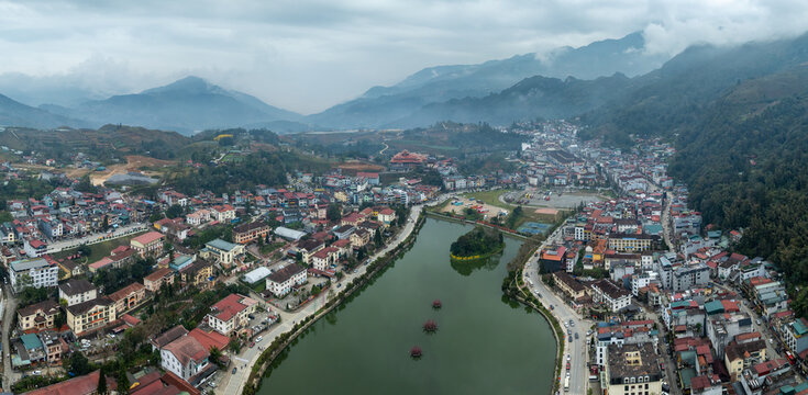 High-angle drone shot of Sapa, Vietnam, featuring its central lake and surrounding buildings, all framed by majestic, cloud-shrouded mountains.
