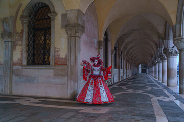 Venetian carnival performer in red and white baroque costume posing under long historic arcade in Venice