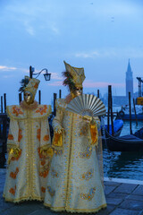 Venetian carnival couple in golden ornate costumes standing by lagoon with gondolas and San Giorgio Maggiore basilica in background