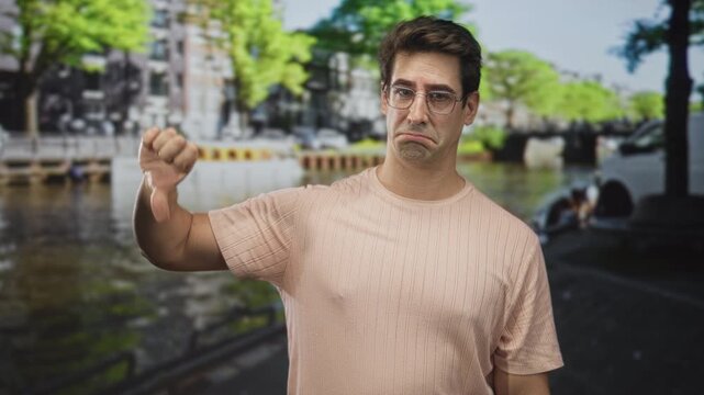 Man points finger up then gives thumbs down on a canal street wearing glasses and a neutral to disappointed expression; disappointment.