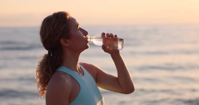 Fit woman drinking water at the beach after running outdoors in the morning. Sporty female relaxing after training against sunrise background. Slow motion