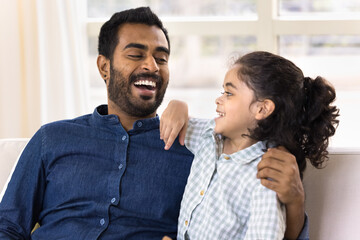 Obraz premium Cute little boy spend pastime with older brother seated on couch. Young Indian dad laughing, talking to preschooler son at home, feel happy, enjoy pleasant communication and time together. Fatherhood