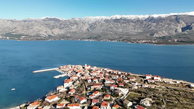 Wide drone orbit around Vinjerac showing red rooftops above blue Adriatic with snowy Velebit peaks and Paklenica canyon visible on a sunny winter day, Dalmatia, Croatia.