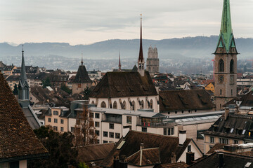 Fototapeta premium A panoramic cityscape of Zurich featuring historic church towers and rooftops under a cloudy sky. The image captures the charm of Switzerland old town with mountains in the background.