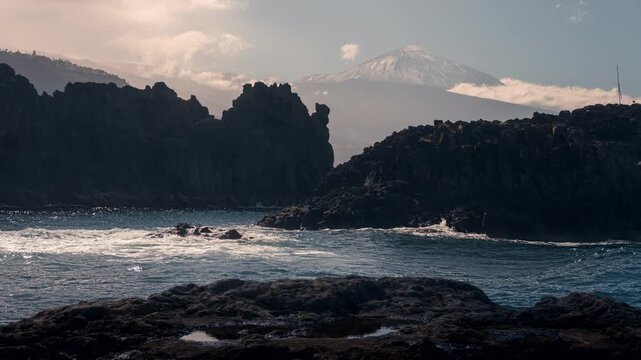 Sunny day timelapse at El Pris on Tenerife&rsquo;s north coast. Waves break against volcanic lava rocks while snow-capped Mount Teide rises in the background under moving clouds.