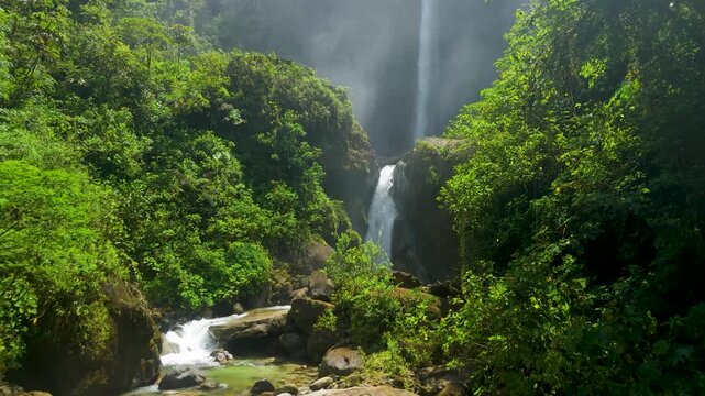 Low altitude aerial shot flying along rocky mountain stream toward magnificent double waterfall Cascada El Rocio Machay hidden in dense tropical rainforest of Banos Ecuador