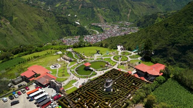 Drone descending over medieval themed Laberinto del Dragon park on mountain slope with panoramic 360 degree views of green Andean valley and Tungurahua Volcano