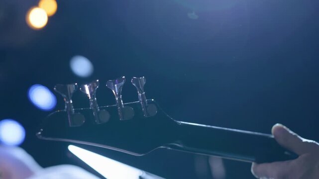 A backlit shot by bright stage lights of the head and tuning pegs of an electric bass with a hand visible playing the instrument during a live performance