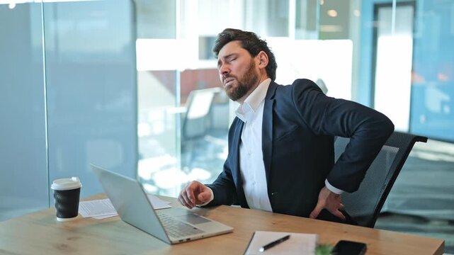 Focused business person feels intense back pain during long hours at office desk. Discomfort strain manifest as chronic posture issues from prolonged laptop use. Suffering and fatigue, seeking relief.