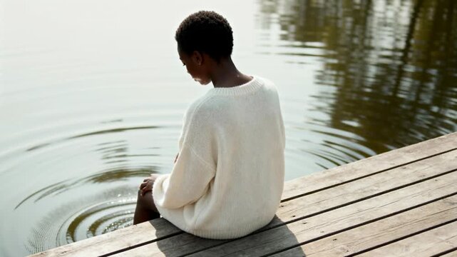 Rear view of a Black woman sitting on a wooden dock by the lake. Person in a white sweater splashing water with feet creating ripples. Solitude and relaxation concept