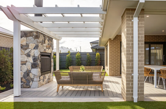 A covered outdoor living space with timber decking, pergola structure, and a stone feature wall fireplace of a contemporary suburban home. Outdoor landscaped backyard living in Australia.