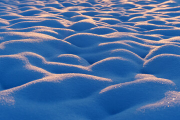 Snow texture forming gentle wavy patterns on a frozen landscape, with morning light casting warm glow on frost patterns and creating blue shadows on cold winter terrain