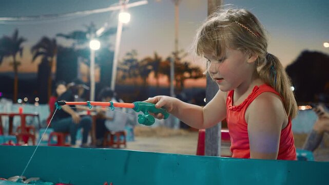 Child enjoys fishing at a night market while people gather around tables in the background during sunset hours
