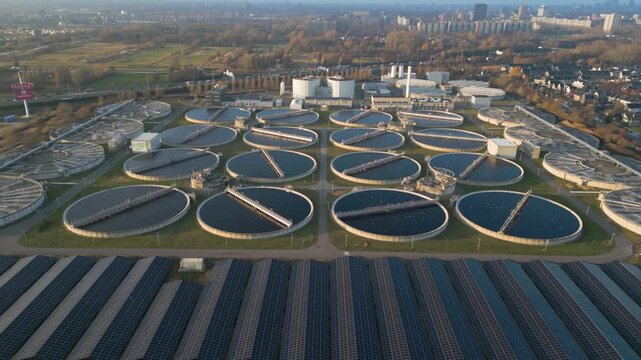 Drone approach shot flying toward large operational wastewater clarifier basins at the Harnaschpolder treatment plant in the Netherlands with sunlight reflections across water surfaces.