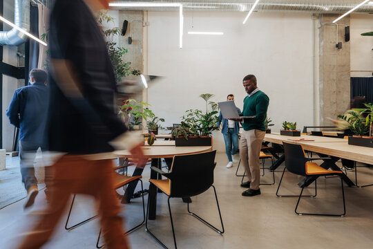 African American man with laptop in a modern Madrid coworking space as coworkers move around