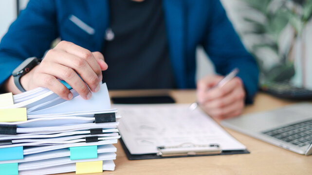 Business professional reviewing and organizing stack of documents with laptop. Concept of paperwork management, administrative workload, compliance, audit process, and office documentation.