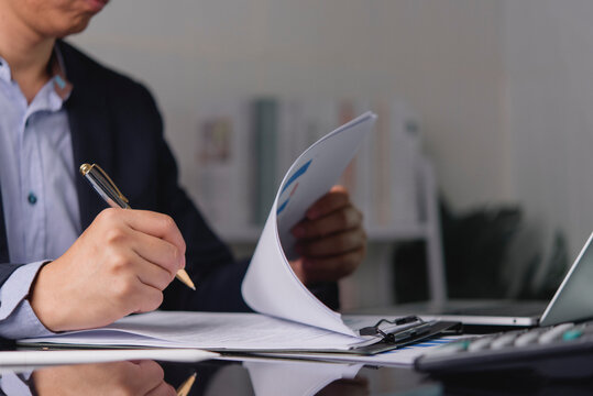 Close up businessman hand holding pen signing contract paper document. Business man working with financial report and calculator at office desk, reviewing loan agreement or tax form.